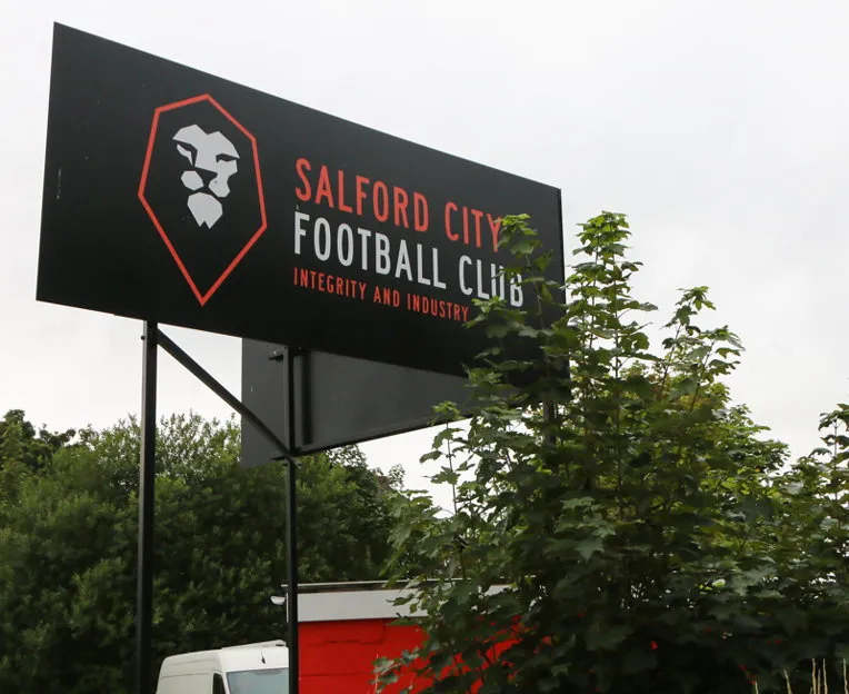 Biển hiệu lớn mang tên Salford City FC màu đỏ trắng nổi bật, tự hào đứng sừng sững bên ngoài sân Moor Lane, là điểm check-in không thể thiếu cho những ai muốn chứng kiến 'đứa con tinh thần' của Giggs, Scholes và bộ tứ huyền thoại.