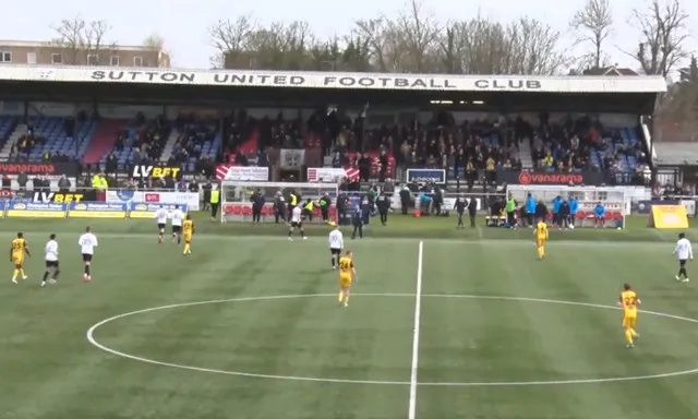 Cảnh trận đấu giữa Sutton United và Torquay United tại Gander Green Lane, thể hiện không khí sôi động trên sân